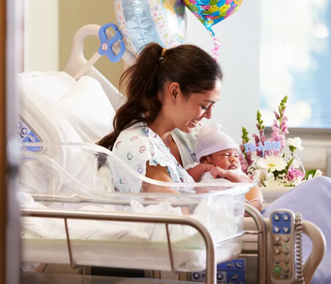 New mom holding baby in hospital bed with flowers and balloons – new baby flower delivery